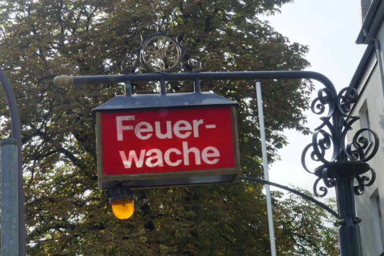 Ein rotes, rechteckiges Schild mit der weißen Aufschrift „Feuerwache“ hängt an einer verzierten Straßenlaterne vor herbstlichen Bäumen.
