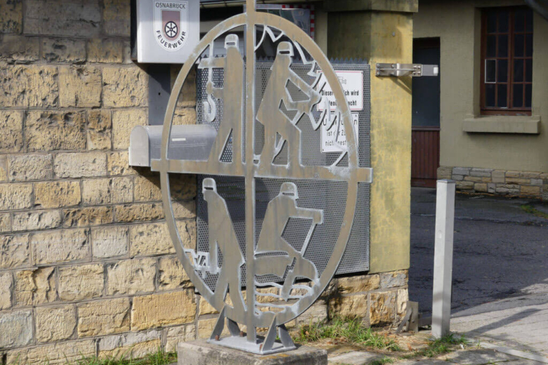 Eine runde, metallische Skulptur vor einer Natursteinwand zeigt vier stilisierte Feuerwehrleute bei verschiedenen Einsatztätigkeiten. Darüber befindet sich das Wappen der Feuerwehr Osnabrück.
