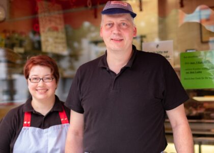 Eine lächelnde Frau mit Schürze und ein Mann mit Kappe stehen vor den Schaufenstern einer Fleischerei.