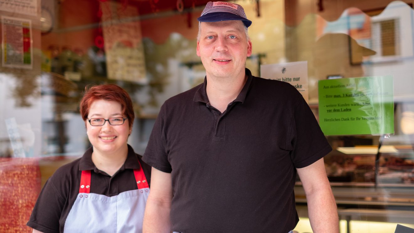 Eine lächelnde Frau mit Schürze und ein Mann mit Kappe stehen vor den Schaufenstern einer Fleischerei.