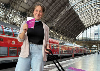 Eine lächelnde Franziska Gröne steht mit einem Koffer auf einem Bahnhofsbahnsteig und hält eine kleine, pinke Broschüre in der Hand. Im Hintergrund ist ein roter Doppelstockzug zu sehen.