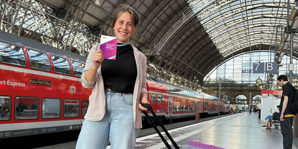 Eine lächelnde Franziska Gröne steht mit einem Koffer auf einem Bahnhofsbahnsteig und hält eine kleine, pinke Broschüre in der Hand. Im Hintergrund ist ein roter Doppelstockzug zu sehen.