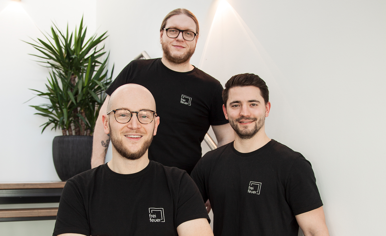 Drei Männer in schwarzen T-Shirts mit Firmenlogo stehen lächelnd vor einer hellen Wand und einer Treppe.