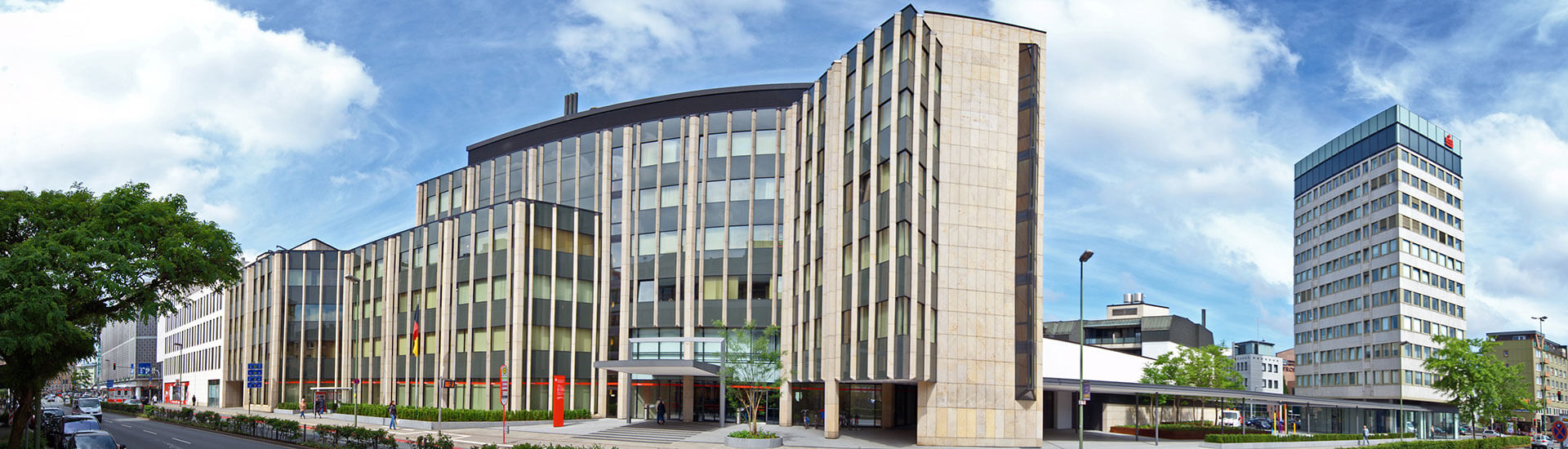 Panoramaaufnahme des modernen Hauptgebäudes der Sparkasse Osnabrück mit hellem Stein, Glasfronten und Hochhaus unter leicht bewölktem Himmel.
