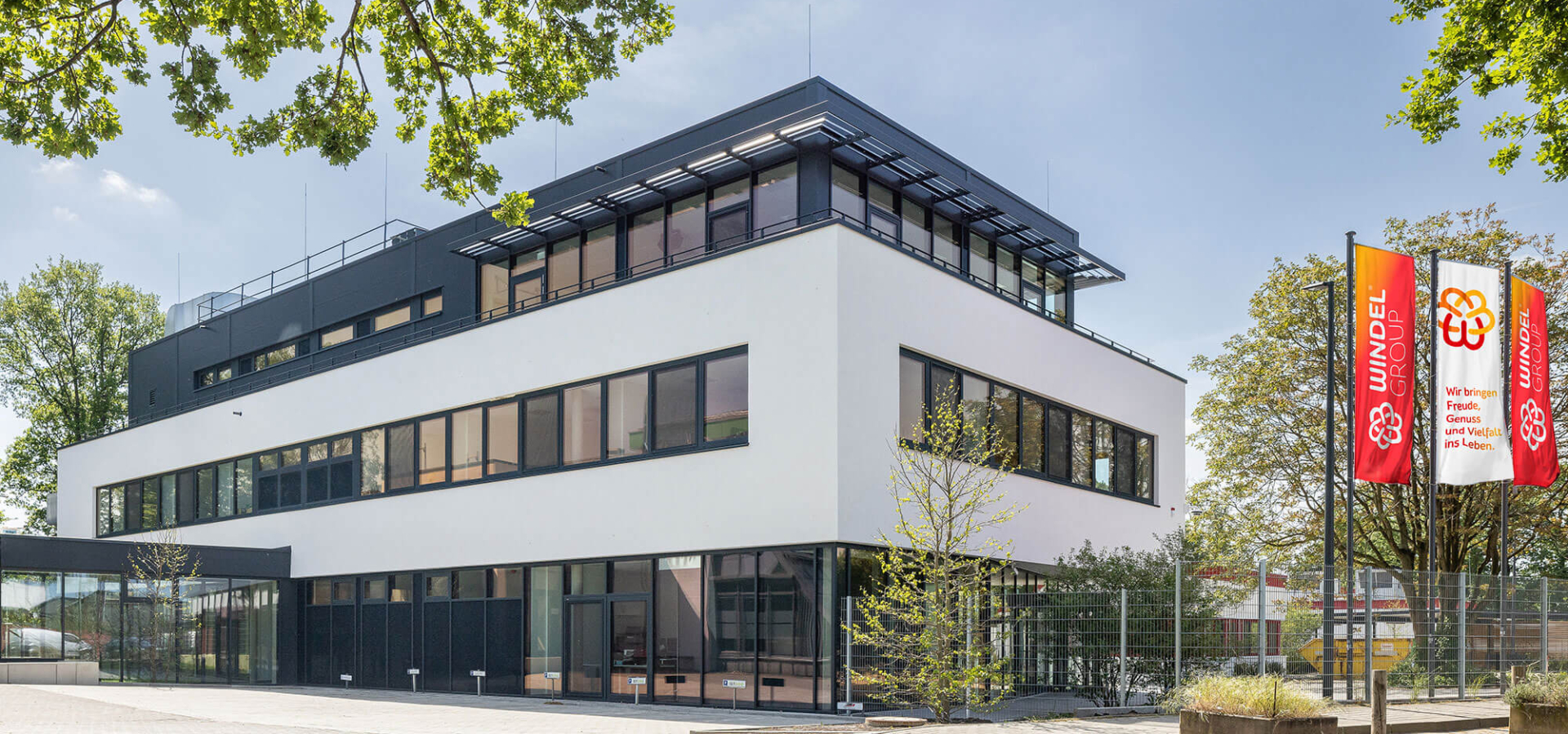 Dreigeschossiges, modernes Firmengebäude der Windel Group mit weißer Fassade, Glaselementen und rot-gelben Fahnen.