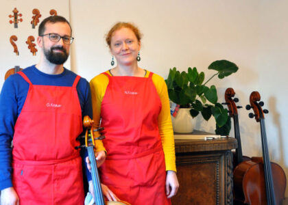 Zwei Personen in roten Arbeitsschürzen stehen lächelnd nebeneinander in einer Geigenbauwerkstatt, umgeben von mehreren Streichinstrumenten wie Geigen und Celli.