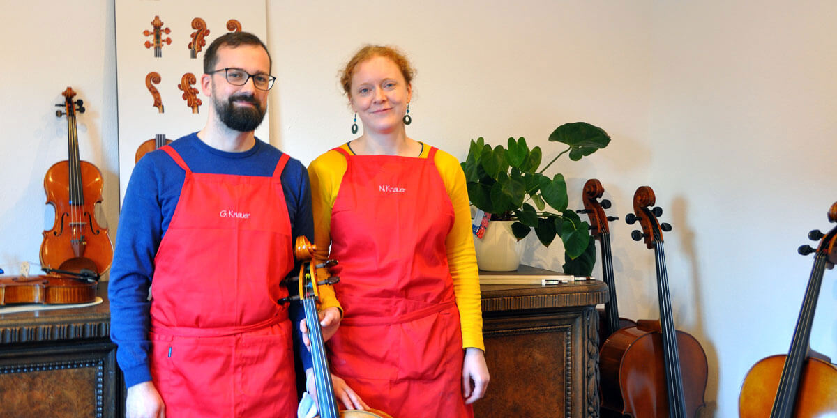 Zwei Personen in roten Arbeitsschürzen stehen lächelnd nebeneinander in einer Geigenbauwerkstatt, umgeben von mehreren Streichinstrumenten wie Geigen und Celli.