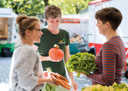 Drei Personen unterhalten sich lächelnd auf einem Wochenmarkt mit Kürbis, Karotten und einem Salat im Arm.