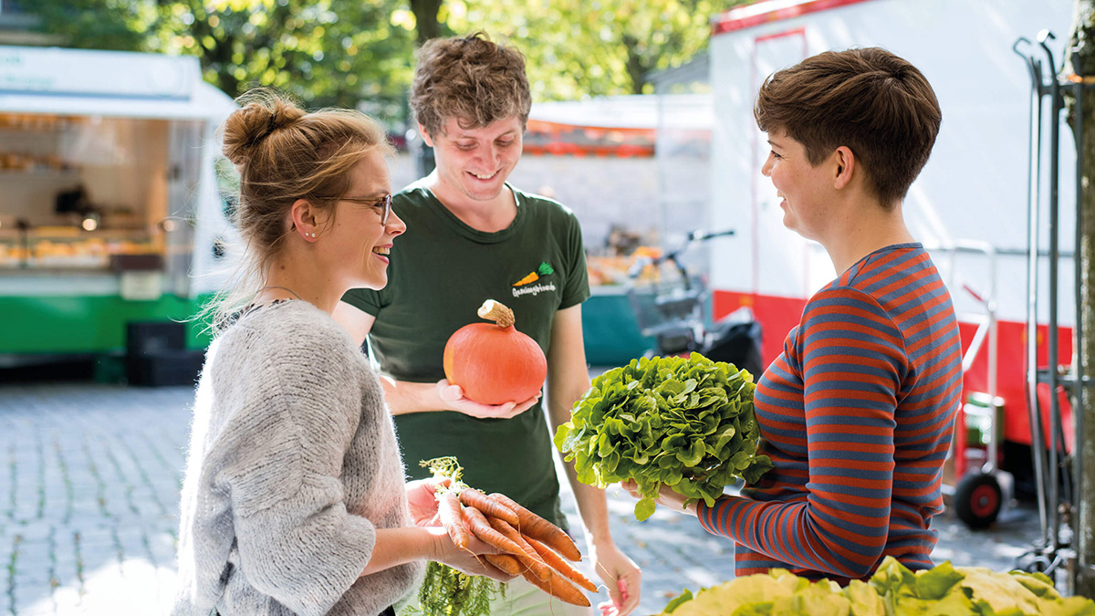 Drei Personen unterhalten sich lächelnd auf einem Wochenmarkt mit Kürbis, Karotten und einem Salat im Arm.