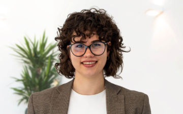Porträt von Merle Meyer mit dunklen Locken, Brille und braunem Sakko vor hellem Hintergrund.