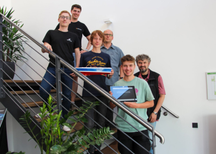 Eine Gruppe von sechs Männern steht auf einer Treppe im Innenbereich und präsentiert ein Schiffsmodell sowie ein Tablet.
