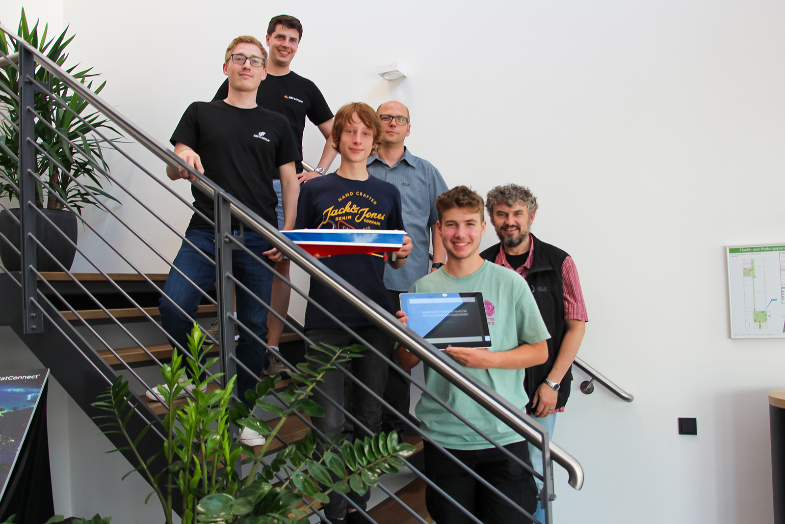 Eine Gruppe von sechs Männern steht auf einer Treppe im Innenbereich und präsentiert ein Schiffsmodell sowie ein Tablet.
