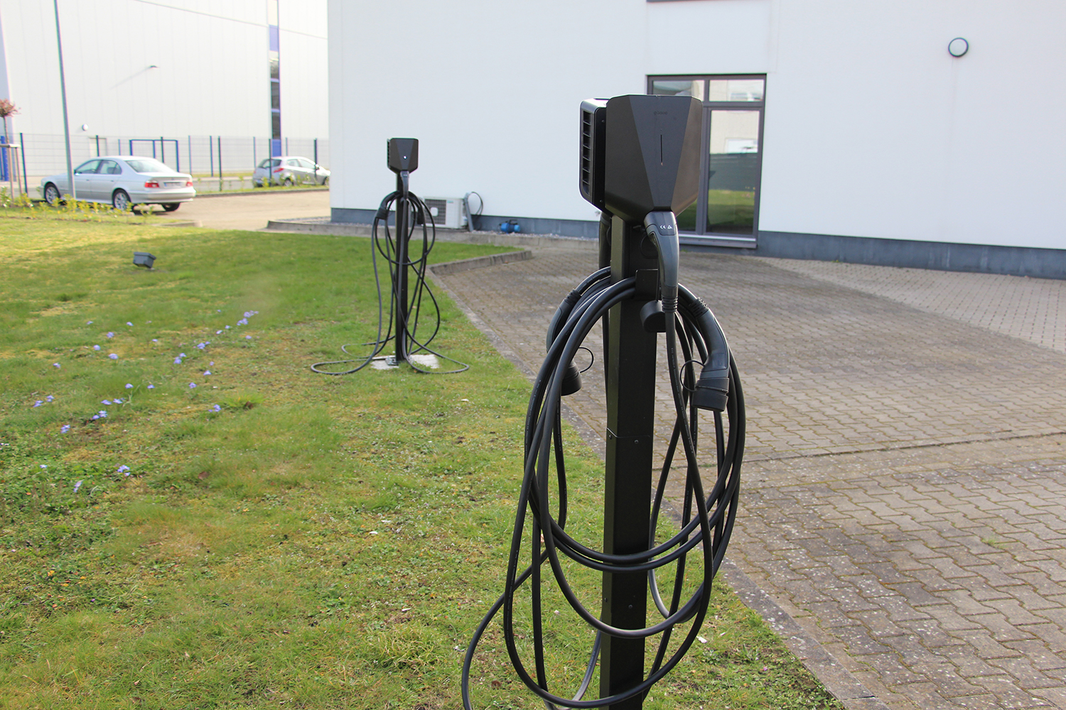 Zwei schwarze E-Ladesäulen mit aufgewickelten Kabeln stehen auf dem Außengelände eines Firmengebäudes.