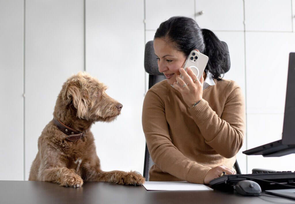 Frau H. Nezir sitzt am Schreibtisch, telefoniert mit einem Smartphone und blickt dabei lächelnd auf einen braunen Hund, der seine Pfote auf den Tisch legt.