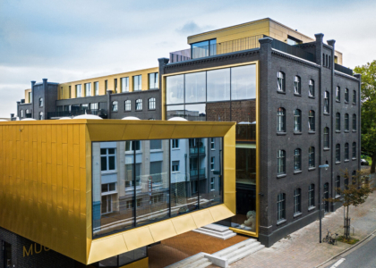 Modernes Gebäude mit dunkler Klinkerfassade und einem markanten, goldfarbenen Anbau mit großen Fenstern.