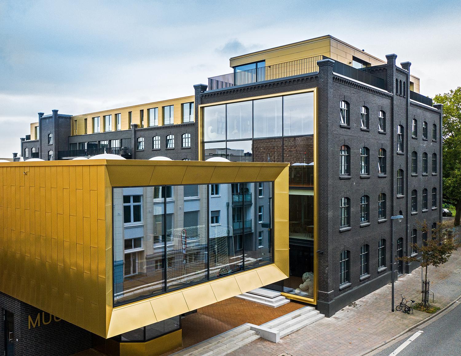 Modernes Gebäude mit dunkler Klinkerfassade und einem markanten, goldfarbenen Anbau mit großen Fenstern.