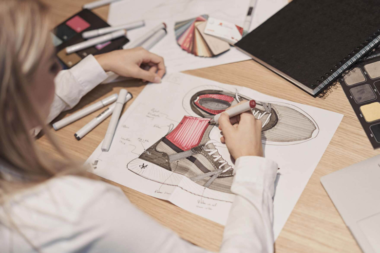 Person zeichnet mit Stiften eine Skizze eines Sneakers auf Papier an einem Holztisch