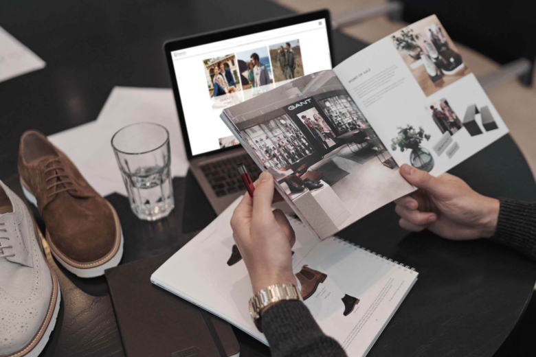 Hände blättern in einem Produktkatalog neben einem Laptop und Schuhmustern auf dem Tisch