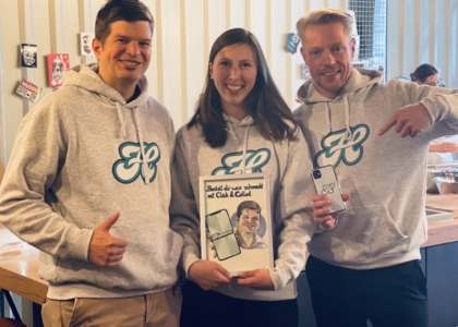 Drei Personen in grauen Marken-Hoodies lächeln; die Frau in der Mitte hält ein Bildrahmen-Schild mit der Aufschrift „Click & Collect“
