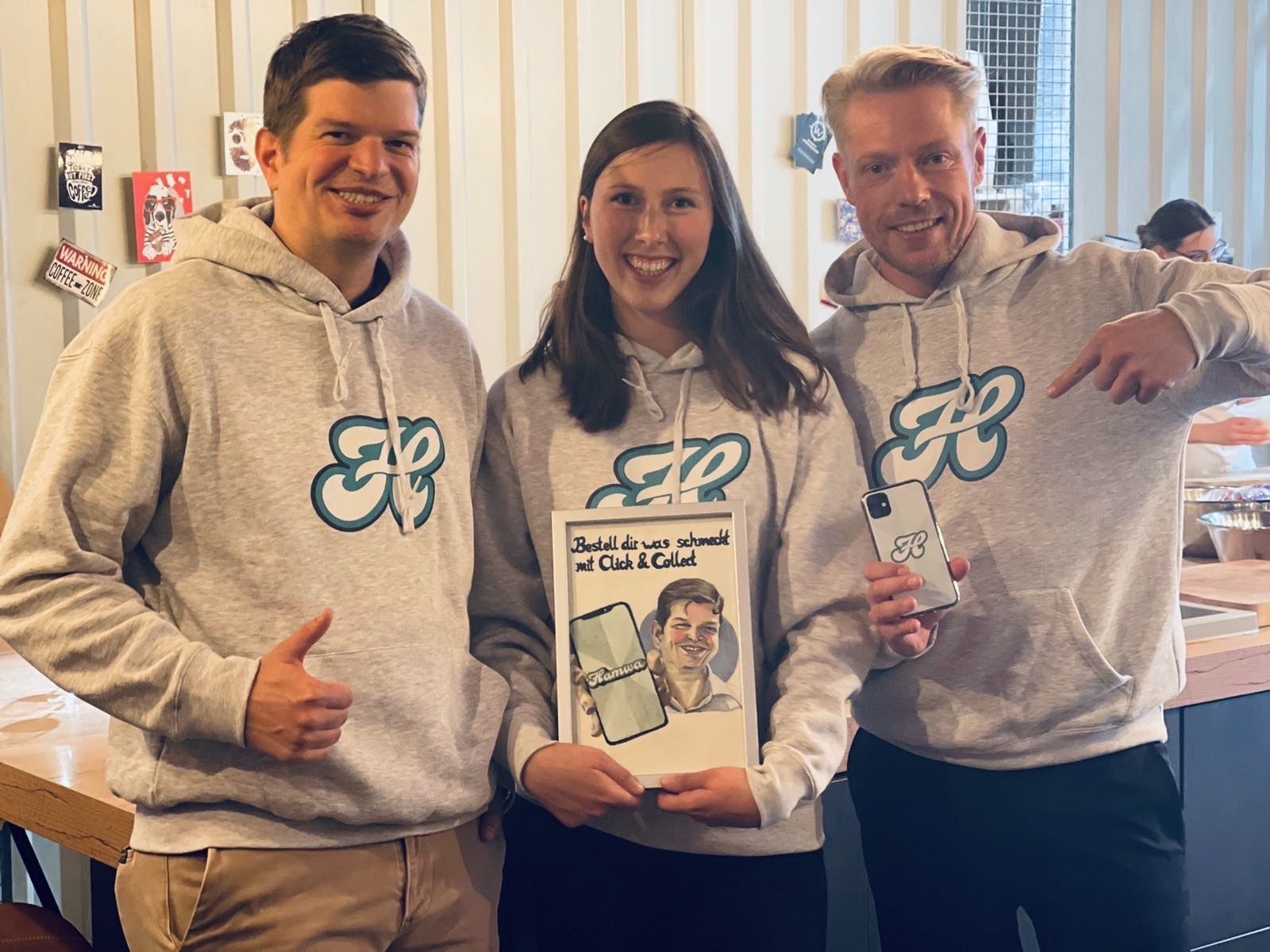 Drei Personen in grauen Marken-Hoodies lächeln; die Frau in der Mitte hält ein Bildrahmen-Schild mit der Aufschrift „Click & Collect“