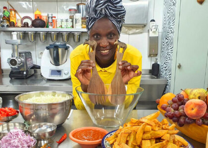 Helen Nintemann mit gelbem Oberteil lächelt in einer Küche; sie hält zwei hölzerne Salatlöffel mit Giraffen-Schnitzerei vor eine Glasschüssel, im Vordergrund steht frisches Gemüse und Obst.