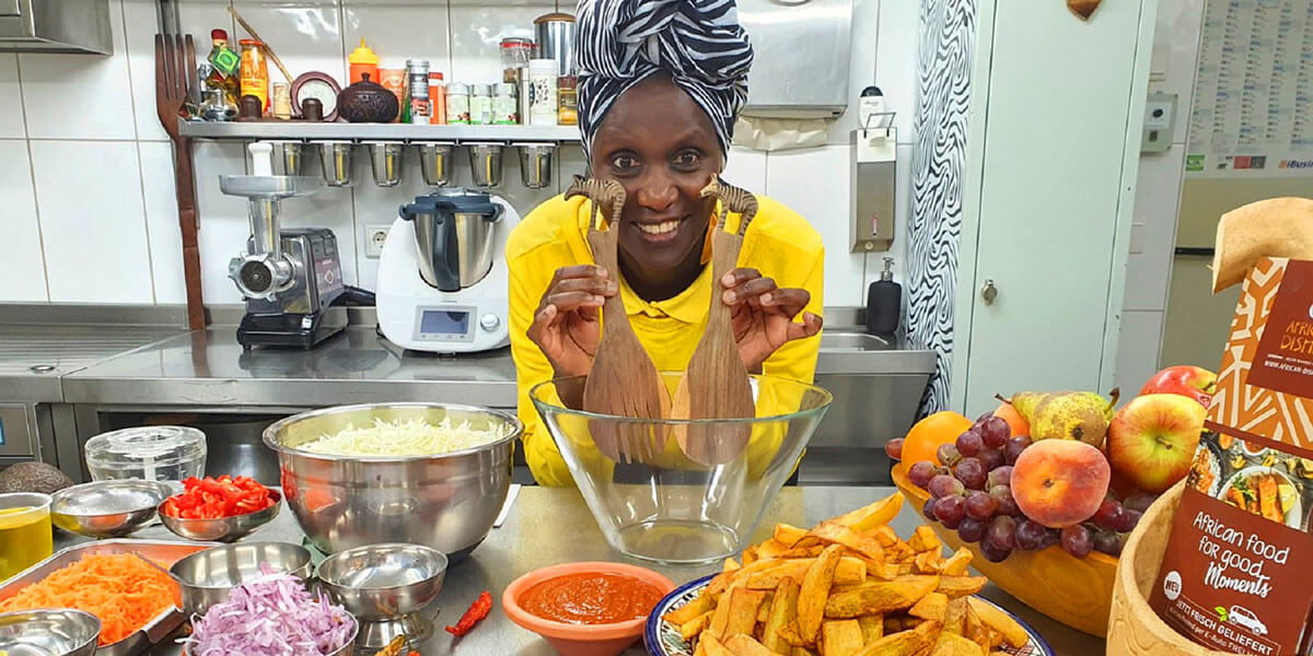 Helen Nintemann mit gelbem Oberteil lächelt in einer Küche; sie hält zwei hölzerne Salatlöffel mit Giraffen-Schnitzerei vor eine Glasschüssel, im Vordergrund steht frisches Gemüse und Obst.