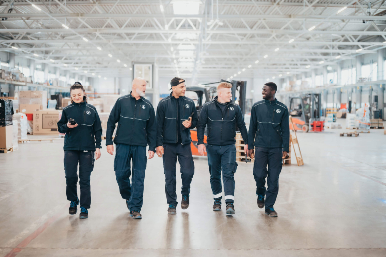 Fünf Personen in dunkler Hellmann-Arbeitskleidung gehen nebeneinander durch eine Logistikhalle.