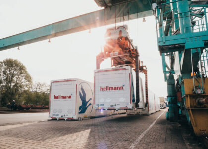 Ein großer Kran verlädt einen weißen Hellmann-Frachtcontainer an einem Güterbahnhof bei Gegenlicht.