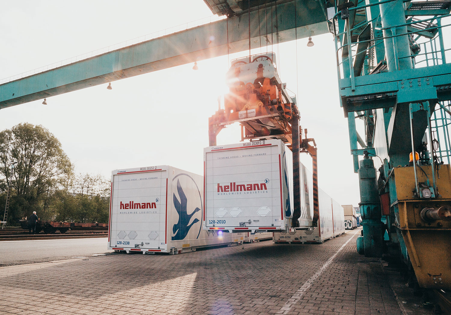 Ein großer Kran verlädt einen weißen Hellmann-Frachtcontainer an einem Güterbahnhof bei Gegenlicht.