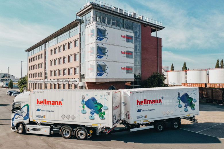 Ein weißer Hellmann-LKW mit Anhänger steht vor einem modernen Firmengebäude.