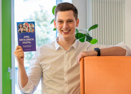 Lächelnder junger Mann im weißen Hemd hält das Buch „JUNG. ERFOLGREICH. DIGITAL.“ in die Kamera und lehnt an einem orangenen Sessel.