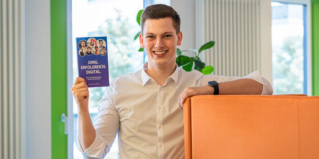 Lächelnder junger Mann im weißen Hemd hält das Buch „JUNG. ERFOLGREICH. DIGITAL.“ in die Kamera und lehnt an einem orangenen Sessel.