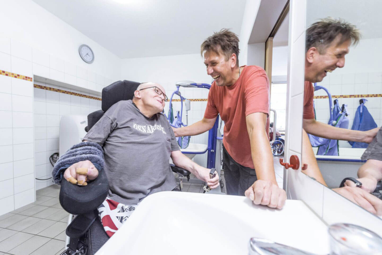 Mann im Rollstuhl lacht gemeinsam mit einem Pfleger im Badezimmer.