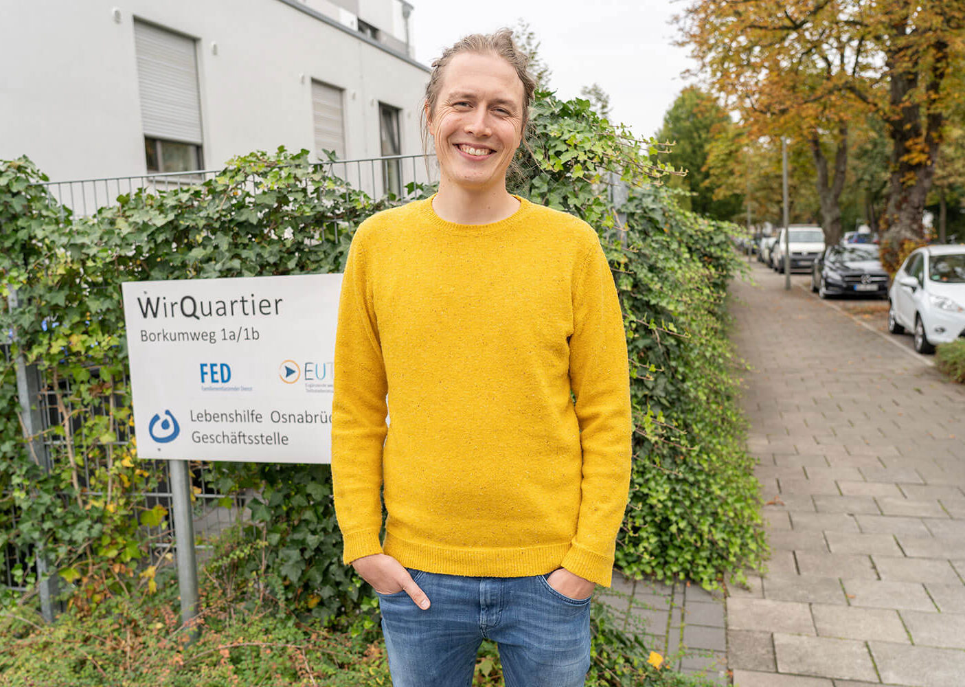 Hendrik de Jong steht lächelnd im gelben Pullover und Jeans auf einem Gehweg neben einem Schild der Lebenshilfe Osnabrück Geschäftsstelle.