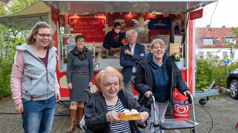 Eine Gruppe von Menschen lächelt vor einem roten Verkaufsstand; eine Frau im Rollstuhl präsentiert stolz eine frische Waffel.