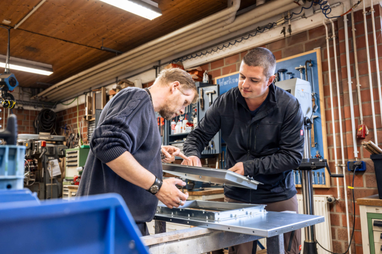 Zwei Männer prüfen gemeinsam ein Metallbauteil in einer Werkstatt.