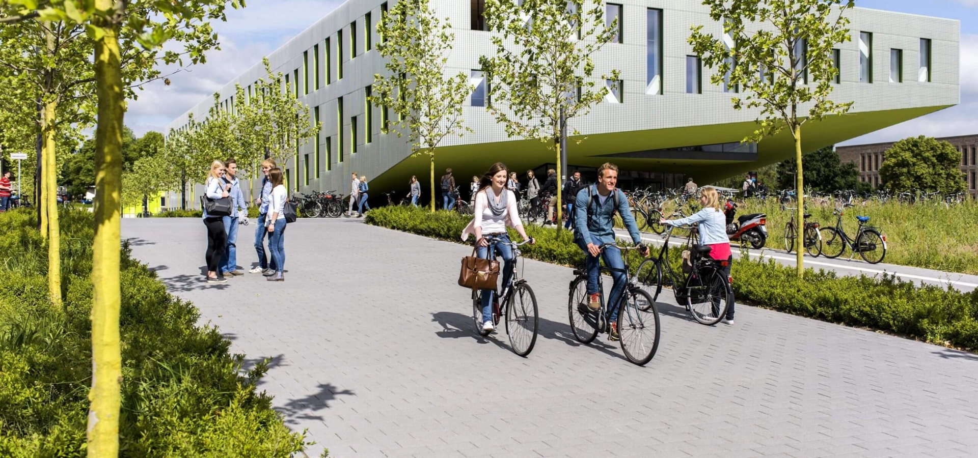 Studierende fahren auf Fahrrädern an einem modernen Campusgebäude mit hellgrüner Fassade und jungen Bäumen vorbei.