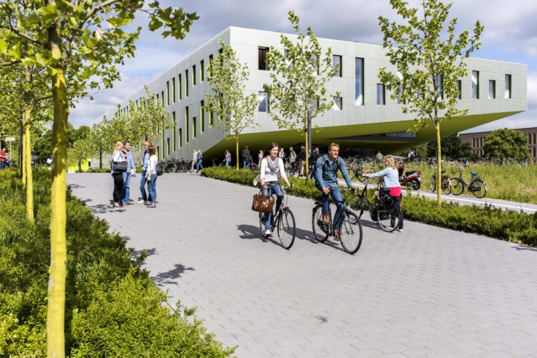 Studierende fahren auf Fahrrädern an einem modernen Campusgebäude mit hellgrüner Fassade und jungen Bäumen vorbei.