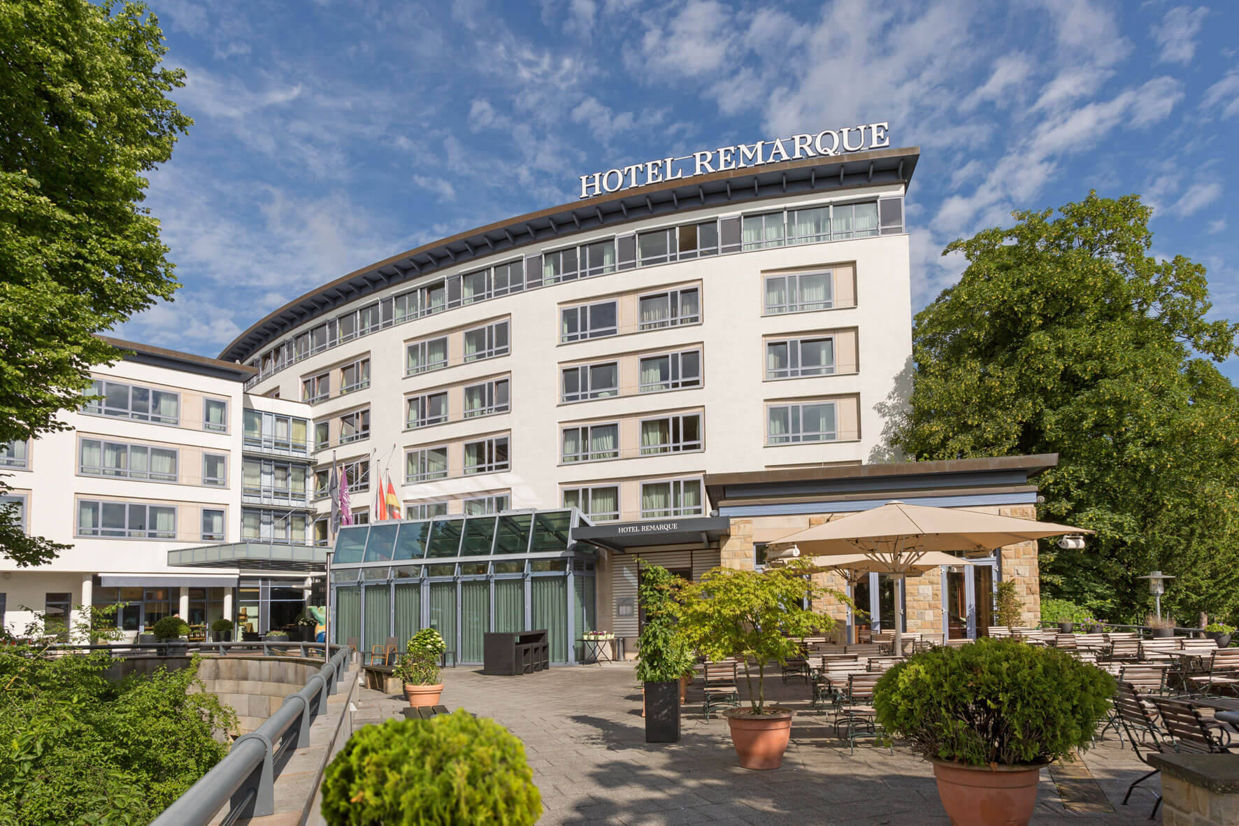 Außenansicht des mehrstöckigen Hotel Remarque mit Terrasse, Sonnenschirmen und Bepflanzung bei blauem Himmel.