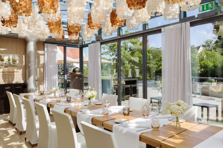 Festlich gedeckte lange Holztafel mit weißen Stühlen in einem lichtdurchfluteten Wintergarten mit Muschelleuchten.