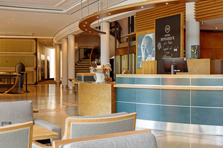 Moderner Empfangsbereich des Hotels mit blau-holzfarbenem Rezeptionstresen, Sitzgelegenheiten und Treppe im Hintergrund.
