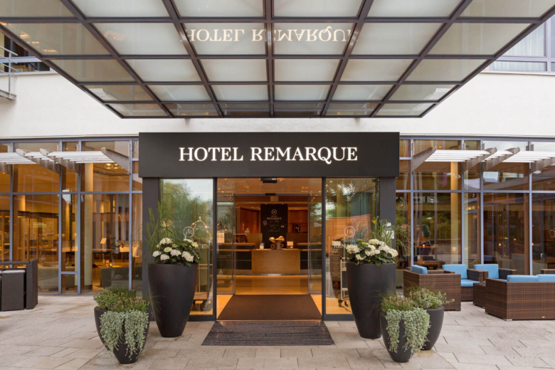 Haupteingang des Hotel Remarque mit Glasschiebetür, Vordach, Schriftzug und großen Pflanzkübeln im Vordergrund.