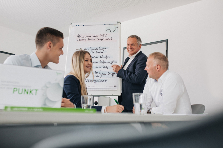Vier Personen sitzen und stehen bei Besprechung um Tisch mit Flipchart in hellem Raum.