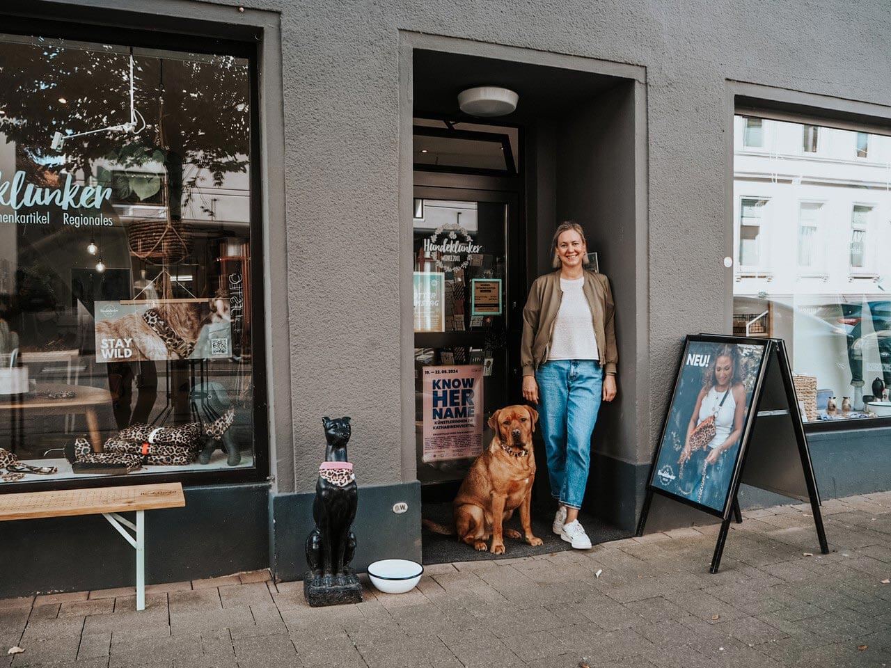 Eine Frau und ein großer brauner Hund stehen im Eingang eines Hundeladens, davor sitzt eine schwarze Hundefigur neben einem Napf.