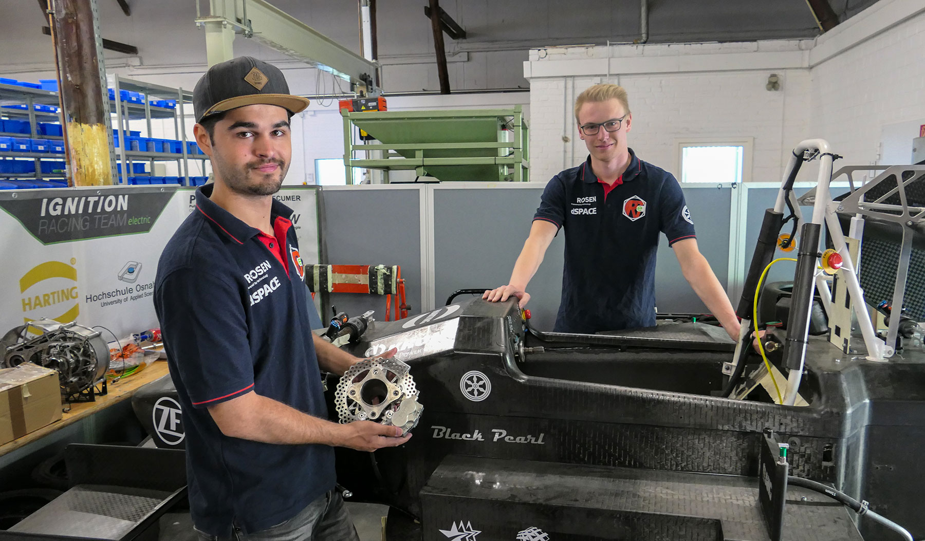 Zwei Männer des Ignition Racing Teams arbeiten in einer Werkstatt an einem schwarzen Formel-Rennwagen und zeigen ein Bauteil.