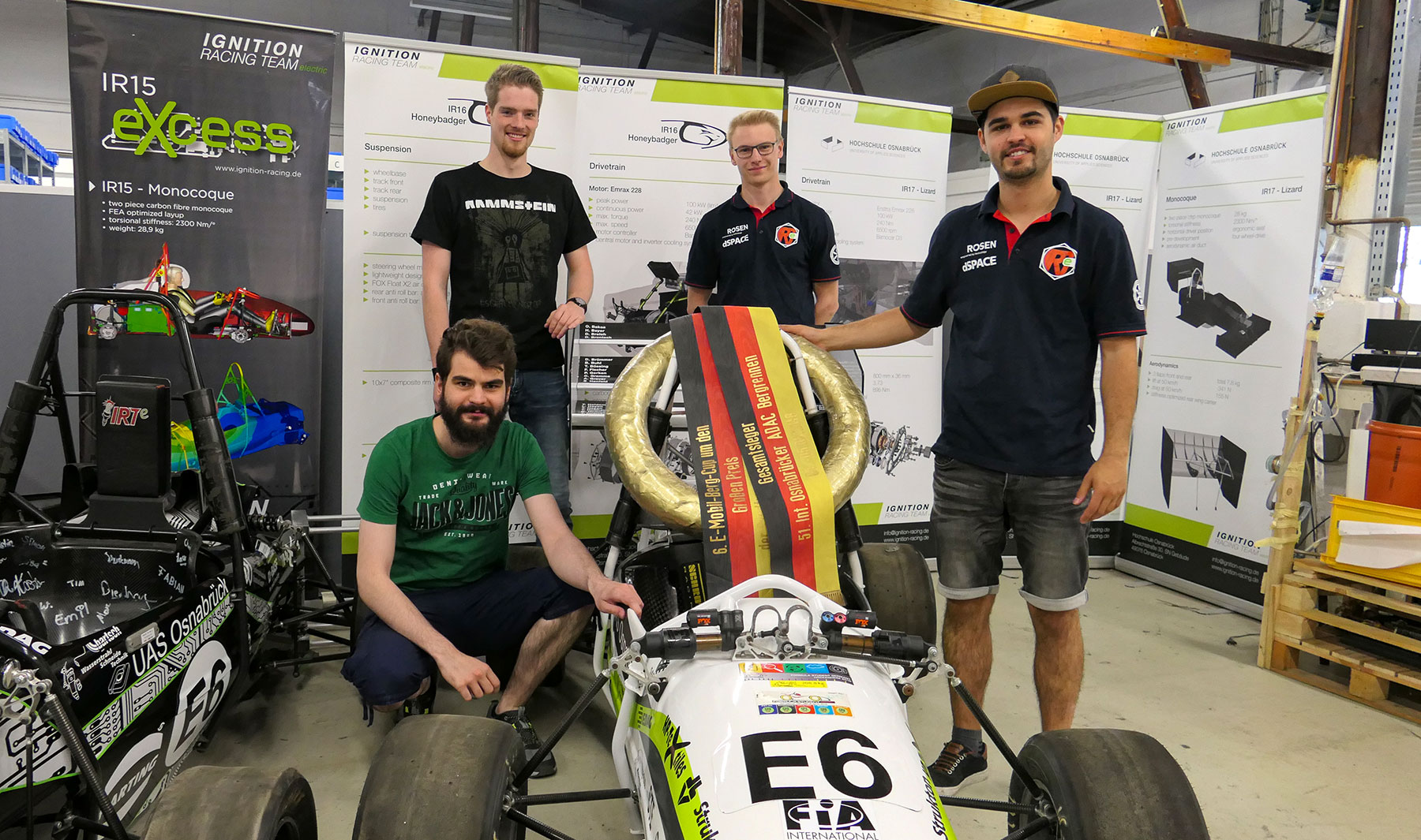 Vier junge Männer des Ignition Racing Teams posieren stolz mit einem Siegerkranz um einen Formel-Rennwagen in einer Werkstatt.