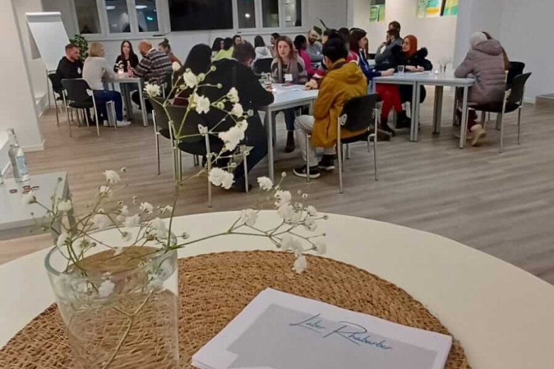 Heller Schulungsraum bei inlingua mit mehreren Lerngruppen an Tischen; im Vordergrund eine Blumenvase und Unterlagen auf einer Tischmatte.