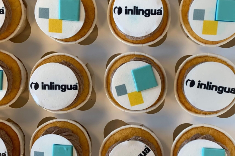 Draufsicht auf mehrere Muffins in einer Schachtel, verziert mit essbarem inlingua-Logo und farbigen Quadraten.