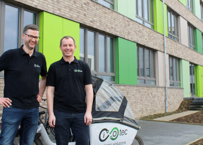 Zwei lächelnde Männer in schwarzen Poloshirts mit iotec-Logo stehen neben einem Lastenrad vor einem modernen Bürogebäude.