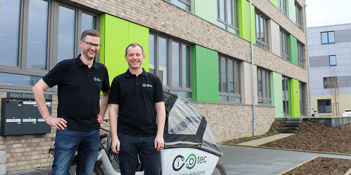 Zwei lächelnde Männer in schwarzen Poloshirts mit iotec-Logo stehen neben einem Lastenrad vor einem modernen Bürogebäude.